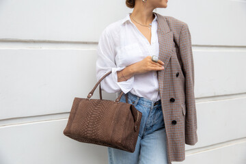 cropped photo of stylish woman in white shirt and oversize jacket with brown bag against wall in...