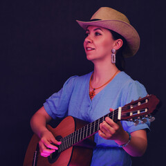 Obraz premium Portrait of a woman with a guitar in her hands on a black studio background. A female musician in a blue dress and a straw hat with an acoustic wooden guitar