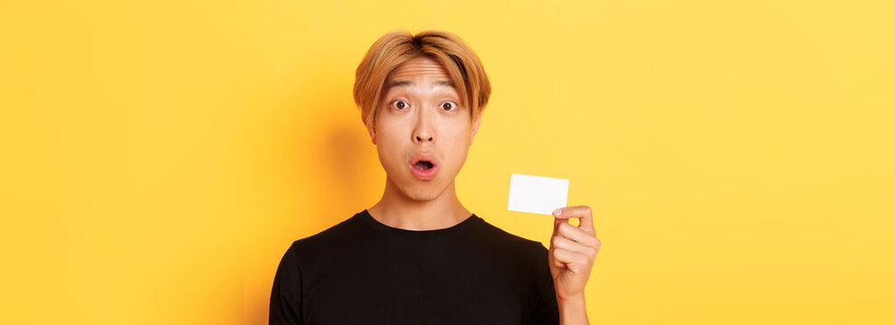 Close-up of surprised gasping asian guy with fair hair, looking amazed and startled as showing credit card, standing yellow background