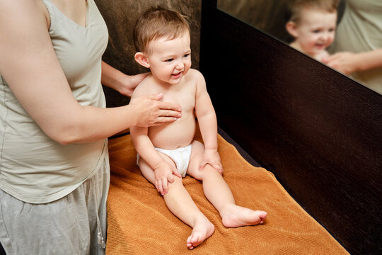 Woman mother giving massage to happy toddler baby sitting on brown towel. Mom massage therapist and smiling child boy in the bathroom. Kid aged one year and two months
