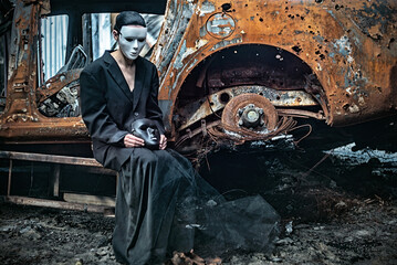 Obraz na płótnie Canvas A girl in a black suit and a white mask sits near a burnt-out car from a Russian army shell hitting the house.