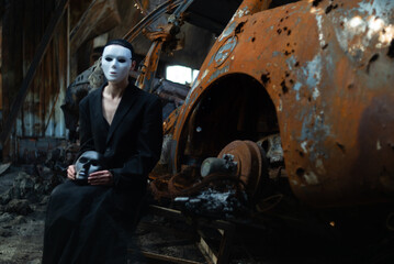 Fototapeta na wymiar A girl in a black suit and a white mask sits near a burnt-out car from a Russian army shell hitting the house.