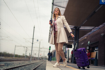 Woman with suitcase waiting for her train on platform of railway station. She is smiling and holding her cup of coffee.