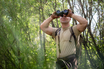 Adventurer man with binoculars in hands on a forest trees background.