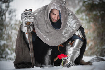 A knight in the armor with the sword and red gerbera flower in the winter snowy forest. Medieval...