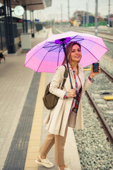 Smiling woman in 30s with backpack and umbrella waiting for her train on platform of railway station. She is holding cup of coffee.