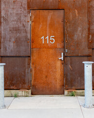 A rusty door with the number 115 is the entrance to a building with a rusty, metal wall. Mooring pilons can been seen in the forefront.