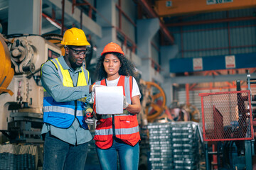 Two professional engineer/worker/technician use clipboard discuss work, walk in steel metal manufacture factory plant industry. Black African American man and woman wear hard hat check quality machine