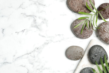 Spa stones with water drops and green leaves on white marble table, flat lay. Space for text