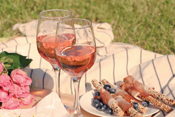 Glasses of delicious rose wine, flowers and food on picnic blanket outdoors