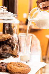 Hand holding a pitcher while pouring milk into a glass tumbler. In the scene there are sweet cookies and a pitcher full of cookies.