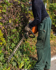 An adult man in overalls cuts bushes with an electric brush cutter in a city park creating a...