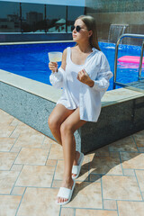 Attractive young woman in a white swimsuit sitting by the pool and drinking a delicious cocktail and sunbathing by the pool during summer vacation