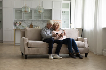 Happy senior family couple enjoying online, Internet communication at home together, sitting on sofa in living room, holding smartphones, studying to use app, service, smiling, talking. Full length