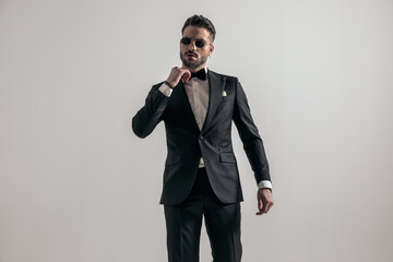 Naklejka premium irresistible man in black tuxedo fixing bowtie while confidently posing