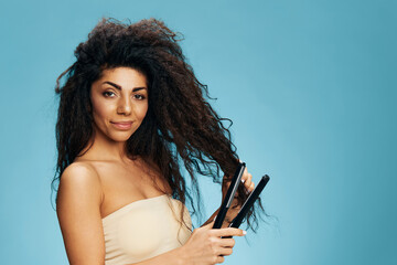 Confident happy Latin curly female using hair straightener, smiling, posing isolated on blue wall...