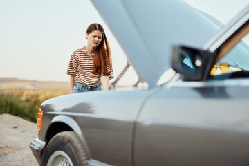 A woman traveler stands by her old car with the hood open, looking for the cause of the car breakdown alone without men on the road in the countryside