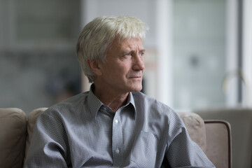 Thoughtful sad senior elder man sitting on home couch, looking away, thinking over health problems, feeling depressed, lonely, frustrated, coping with loss, grief, disease, mental disorder