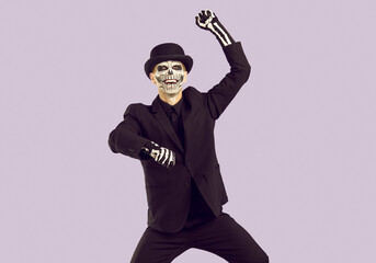 Cheerful man with skeleton makeup on his face having fun dancing at Halloween costume party. Man in black suit, hat and gloves with painted bones is dancing in gangnam style on light lilac background.