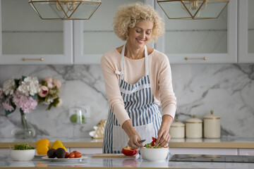 Happy pretty middle aged chef woman slicing fresh vegetables on kitchen table, cooking healthy vegetarian, vegan, raw food diet meal, using organic food ingredients, smiling, laughing