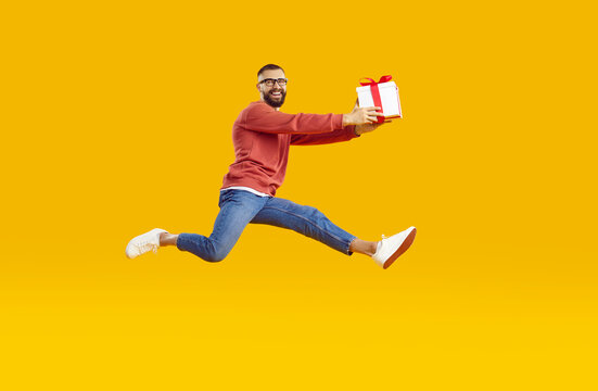 Handsome man in casual clothes and glasses holding gift box and jumping in air isolated on orange yellow background. Happy guy with present running at full speed as he is hurrying to friend's birthday