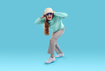 Funny, crazy woman in fashion studio. Excited young girl wearing faux fur bucket hat, sunglasses, mint cotton sweatshirt, grey trousers and white sneakers standing on blue background and screaming