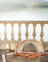 rear view of a woman with a hat and her hands crossed behind her head, sunbathing and resting on a deck chair in front of a lake. vertical format. concept of relaxation and tranquility.