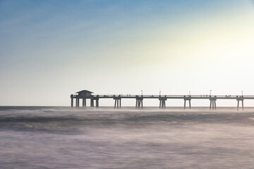 A long exposure photo of a pier on the Gulf of Mexico in Florida in the middle of the day with a blue and golden sky. 