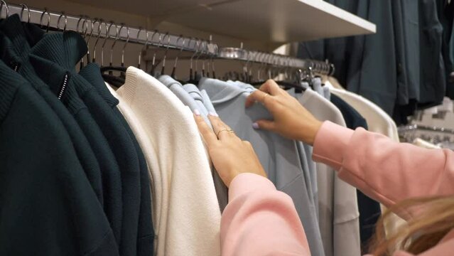 The girl chooses a warm knitted sweater in the store. Women's hands move hangers with clothes. Shopping concept.