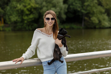 White Caucasian woman holds her pet  short-muzzled dog of breed 'Petit Brabancon Griffon Belge' on hands like a baby on pierce of lake, green trees are on background. Family true friendship concept.