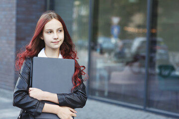 Young girl with closed laptop standing in front of a glass window in the city