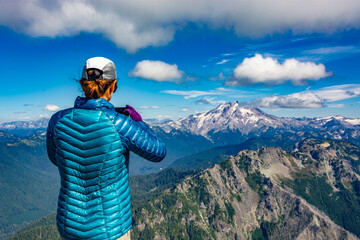 Obraz premium Adventurous athletic female hiker taking a photo with her cellphone of jagged mountains in the in the Pacific Northwest.