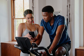 Obraz premium Two young multiracial friends using mobile phone on exercise bike at the gym