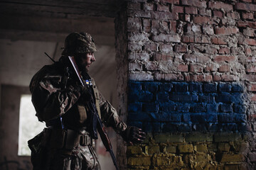 Fototapeta na wymiar A Ukrainian military man with a weapon in his hands stands against the background of the flag of Ukraine. The flag of Ukraine is painted on a brick wall.