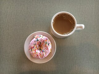A rainbow sprinkle donut on a small white plate next to a mug of coffee