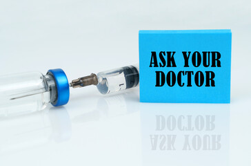 On a white reflective surface there is a syringe with an ampoule and a plate with the inscription - ASK YOUR DOCTOR