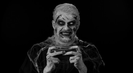 Sinister man with horrible scary Halloween zombie makeup enthusiastically playing racing drive video games on mobile phone. Dead guy with wounded bloody scars face isolated against black background