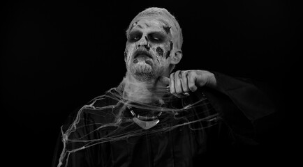 Sinister man Halloween crazy zombie with bloody wounded scars face trying to scare showing killing gesture, runs a finger along his neck isolated on black room. Horror theme of cosplay wounded undead