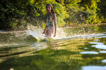 A teenage girl splashing in green water of a river, lake in summer day. Summer vacation fun, joy. Female child 10-12 years at a children's camp. Fun, joy, excitement. Green wave, splashing water.