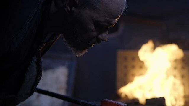 Dark shot of the blacksmith working in the forge striking the glowing metal in the smithy. Hammer on the anvil and sparks in slow motion.