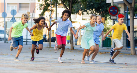 Cheerful active tween boys and girls in colorful clothes running holding hands along summer city street.. - obrazy, fototapety, plakaty