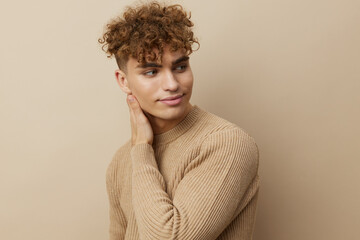 Fototapeta na wymiar a charming young man with curly hair stands on a beige background in a stylish sweater and looks away, relaxed touching his neck with his hand. Horizontal studio photography with empty space