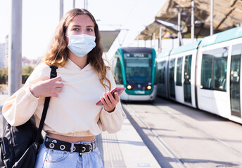 Portrait of a confident girl in a protective mask, standing at a tram stop during a pandemic...