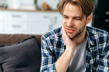 Problems with teeth. Toothache. Upset caucasian guy is sitting on the couch at home, holding his...