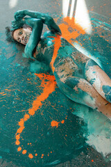 expressive sexy naked woman on the floor in turquoise blue and orange color abstractly painted bodypainting girl on the splashed ground