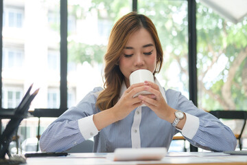 Pleasant young woman enjoying her morning coffee while sitting at her workplace.