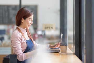 Portrait of a friendly young asian business woman wearing apron and using laptop in her coffee shop, open sign barista SME entrepreneur seller concept