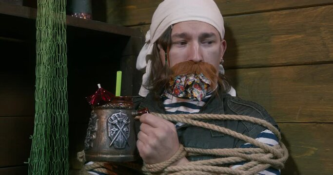 Rowdy sailor tied with rope sits in hold bound with rope, gagged. Pirate after night of revelry wants get drunk. Man with mug in his hands, bandana was taken hostage. Corsair punished for debauchery