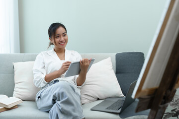 Portrait of an Asian woman using a tablet to paint artwork on a sofa.