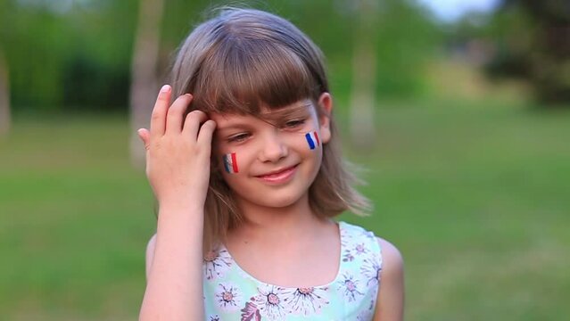 Slow motion of adorable little girl smile at camera with cheeks painted in flag of France. Bastille day on July 14. The day of France. French summer holidays, travel concept.